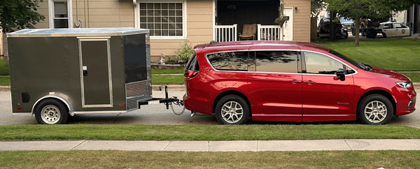Red minivan towing a gray trailer parked on a suburban street next to a grassy lawn.