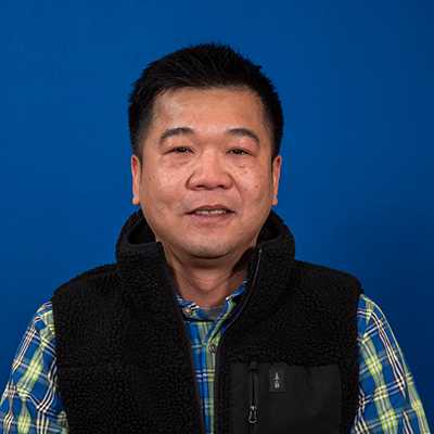 A smiling middle-aged man who has black, short hair wearing a vest over a blue and green plaid shirt.