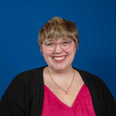 A white woman in her 30s with short blonde hair and cat-eye glasses, in a bright pink blouse smiling broadly at the camera.