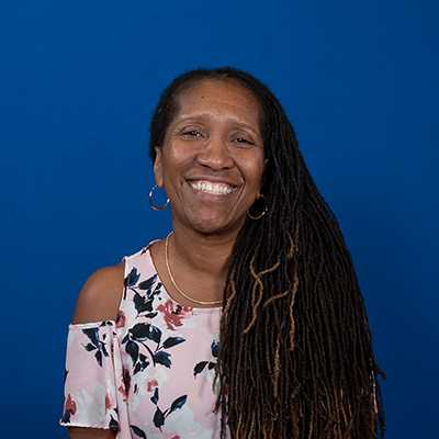 A middle-aged, African American female with long hair in locs, wearing a pink and blue flower print blouse.